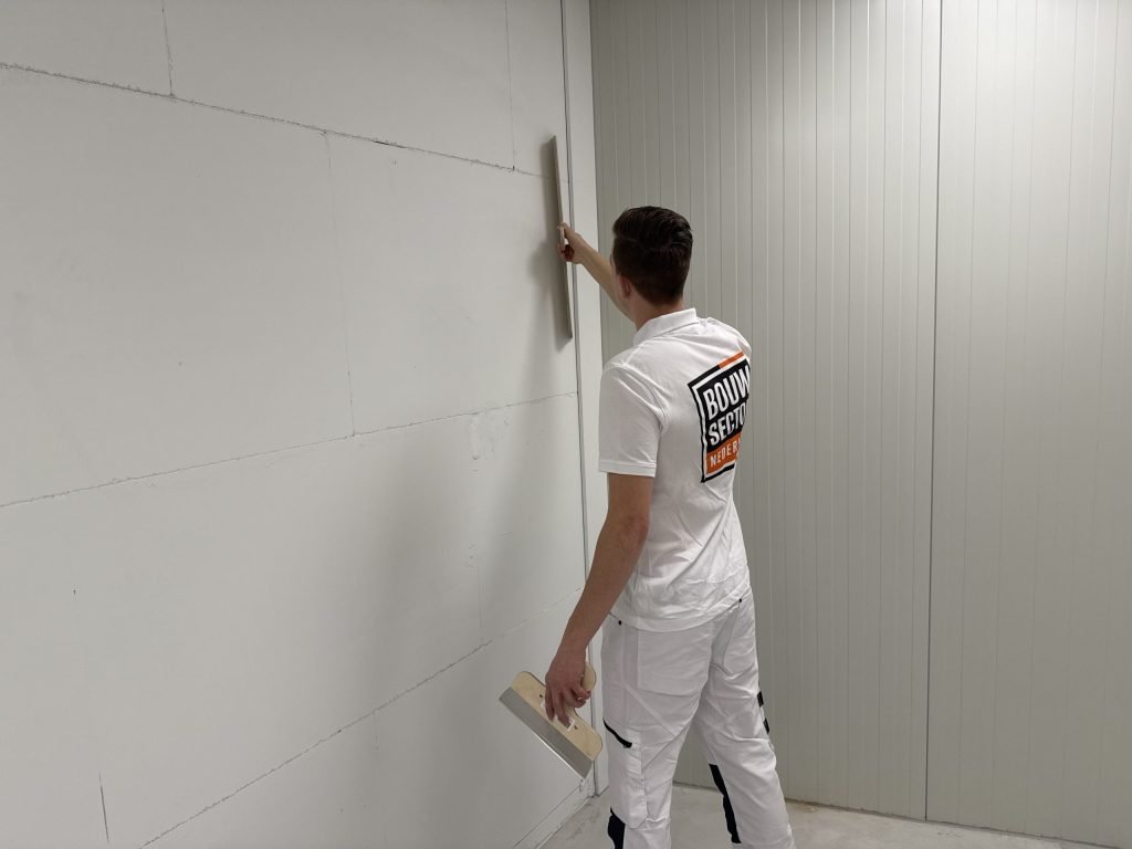 Stukadoor in wit werkpak zet een lange spaan verticaal tegen een wand van gipsblokken, met een pleisterspaan in de andere hand